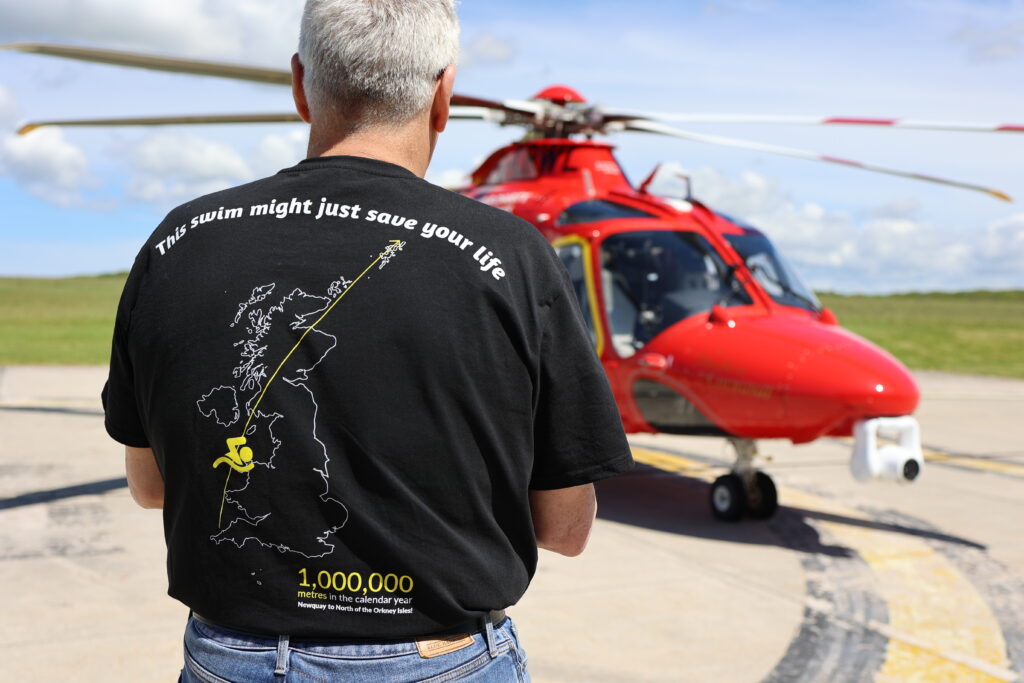 John standing on the heli-landing pad, facing the red helicopter in a black charity t-shirt that says 'this swim might just save your life.'