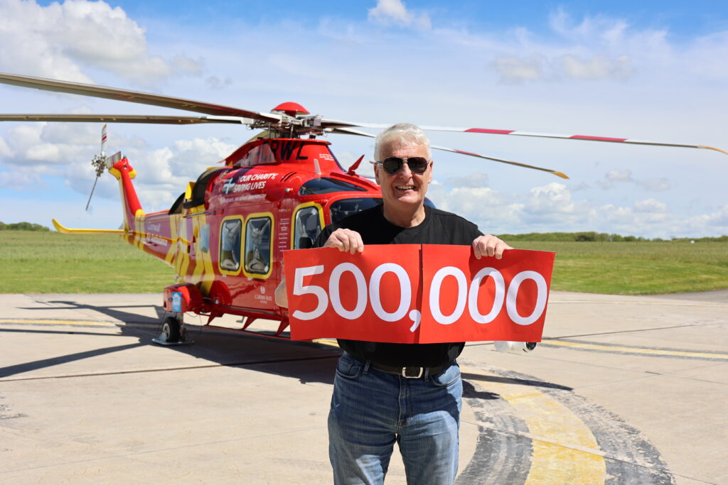 John holding a large red banner than says 500,000 on the heli-pad, with Cornwall Air Ambulance helicopter behind him.