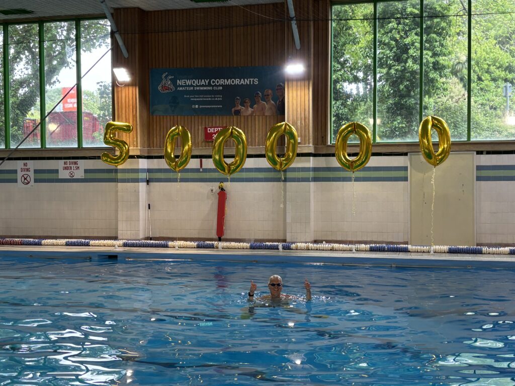 John holding both of his thumbs up in a swimming pool, with golden numbered balloons behind him that spell out 5,00, 000.