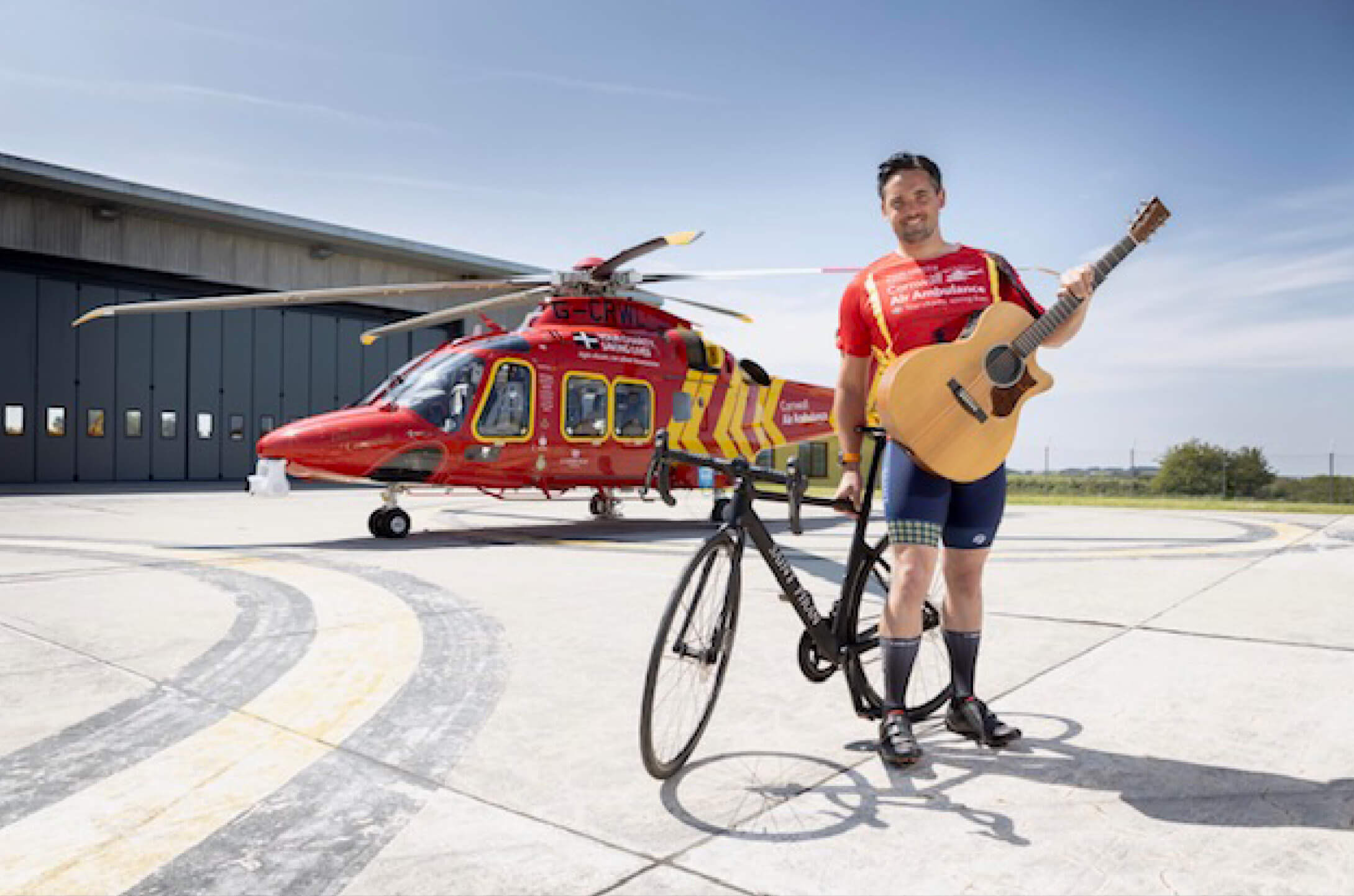 Tom Cary Supporter in front of helicopter with bike and guitar