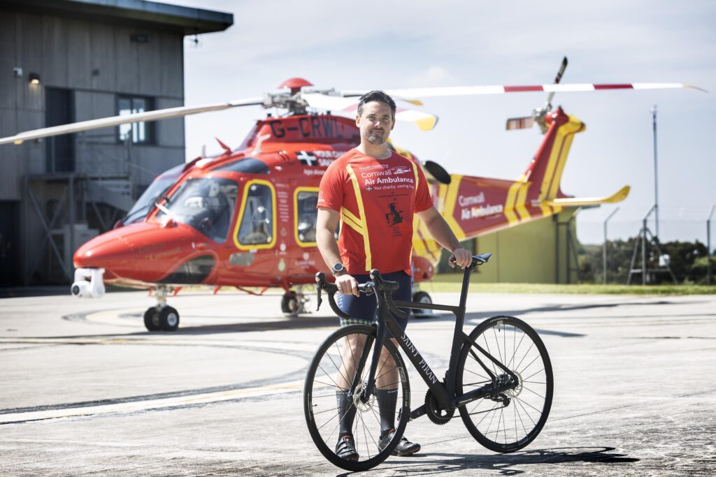 Tom Cary standing with his bike on the heli-pad with the Cornwall Air Ambulance Helicopter behind him.
