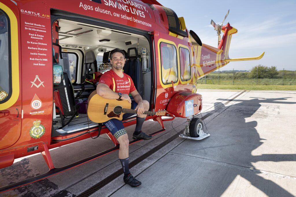 Tom sitting in the side door of Cornwall Air Ambulance helicopter, on the landing pad holding his guitar.